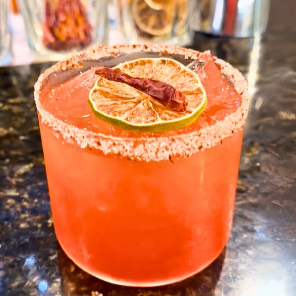 A vibrant red Thai cocktail, garnished with a dried lime slice and a red chili, on a dark counter.