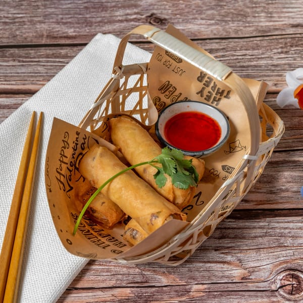 Crispy Thai spring rolls with sweet chili sauce and cilantro in a bamboo basket on a wooden table.