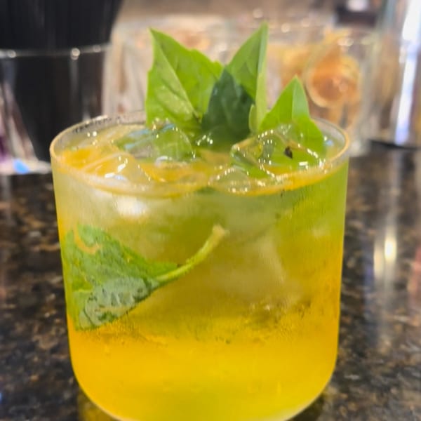 A vibrant yellow Sampannee Thai cocktail with ice and fresh green basil leaves on a dark counter.