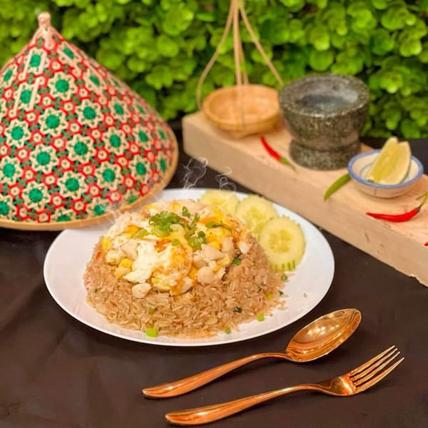 Steaming Thai fried rice with chicken, egg, and cucumber, alongside traditional decor and gold utensils.