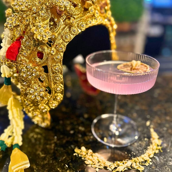 A pink cocktail with a citrus slice next to a golden, jeweled Thai headdress.