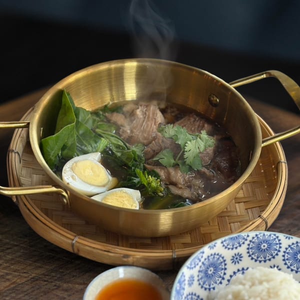 Steaming Thai beef stew with greens and hard-boiled eggs in a brass pot, with rice and sauce.